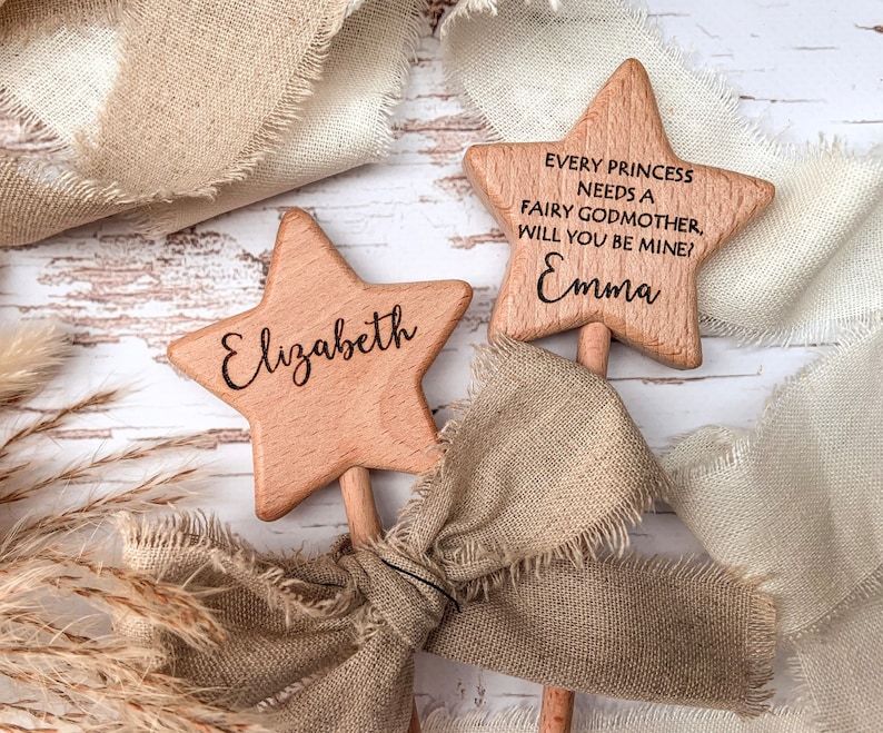 Puede incluir: Dos varitas de madera de color marr&oacute;n claro en forma de estrella con palos. Una varita est&aacute; grabada con el nombre Elizabeth. La otra varita tiene grabado el texto Every princess needs a fairy godmother, will you be mine? y el nombre Emma en negro.