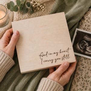 Könnte beinhalten: Eine hellfarbene Holzkiste mit der Inschrift "And in my heart I carry you still" in schwarzer Schrift. Neben der Kiste befindet sich ein Ultraschallbild. Eine brennende Kerze in einem Glashalter und Grünzeug befinden sich im Hintergrund.