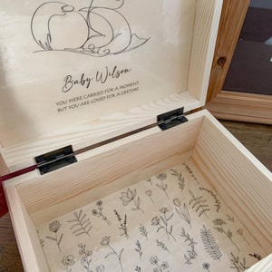 May include: Open wooden keepsake box with a floral patterned interior. The inside lid features a line drawing of a baby with the name "Baby Wilson" and the text "YOU WERE CARRIED FOR A MOMENT, BUT YOU ARE LOVED FOR A LIFETIME."