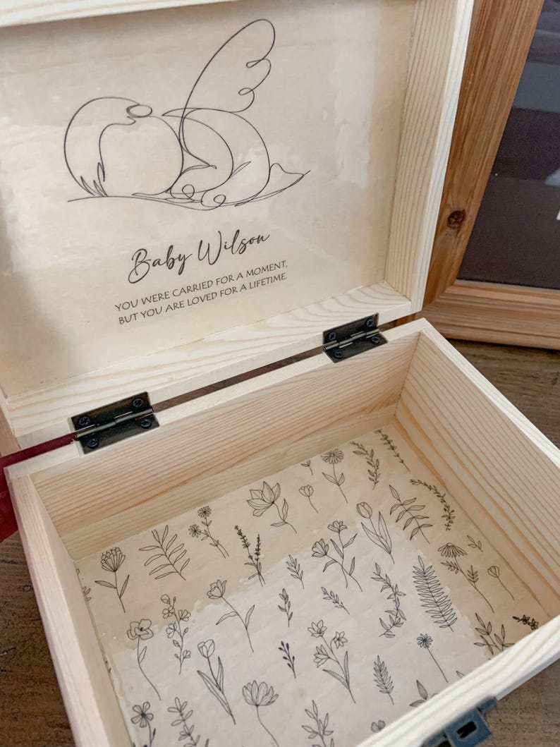 May include: Open wooden keepsake box with a floral patterned interior. The inside lid features a line drawing of a baby with the name "Baby Wilson" and the text "YOU WERE CARRIED FOR A MOMENT, BUT YOU ARE LOVED FOR A LIFETIME."
