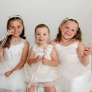 Peut inclure: Trois enfants portant des robes blanches et des bandeaux de perles tiennent des baguettes d'&eacute;toiles en bois avec des rubans roses. Les baguettes portent les noms "Grace", "Nova" et "Lila" &eacute;crits dessus.