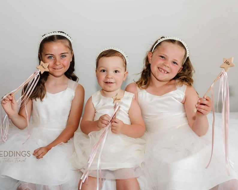 Peut inclure: Trois enfants portant des robes blanches et des bandeaux de perles tiennent des baguettes d'&eacute;toiles en bois avec des rubans roses. Les baguettes portent les noms "Grace", "Nova" et "Lila" &eacute;crits dessus.