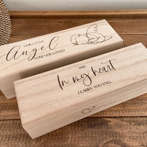 Könnte beinhalten: Zwei rechteckige Holzkisten mit graviertem Text und Illustrationen. Eine Kiste trägt die Aufschrift "OUR LITTLE Angel FOREVER LOVED" mit einer Engel-Illustration. Die andere Kiste trägt "In my heart I CARRY YOU STILL" mit einem Herzsymbol.