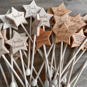 Puede incluir: Una colecci&oacute;n de varitas de madera clara con forma de estrella, cada una unida a un fino palo de madera. Las estrellas est&aacute;n grabadas con varios nombres y t&iacute;tulos como Auntie, Godmother y Flower Girl, junto con breves mensajes personalizados.
