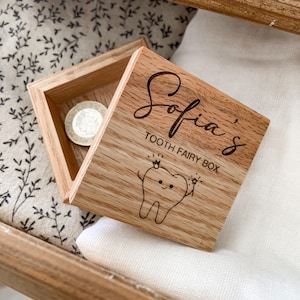 Könnte beinhalten: Eine kleine Holzkiste mit einem Scharnierdeckel. Die Kiste ist mit dem Text "Sofia's Tooth Fairy Box" und einem Cartoon-Zahn mit Krone und Zauberstab graviert. Die Kiste ist geöffnet und eine Münze ist im Inneren sichtbar.