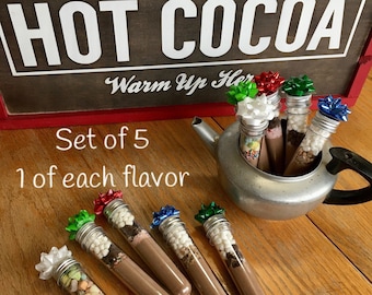 May include: A set of five hot cocoa mixes in clear tubes, each topped with a colorful bow. The tubes are filled with cocoa powder and toppings. A sign in the background reads "Old Fashioned Genuine Hot Cocoa."