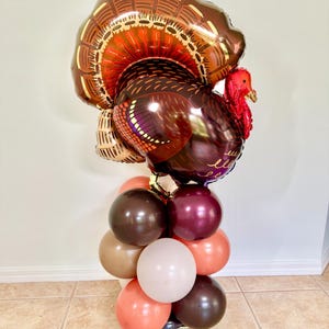 May include: A large brown and red foil balloon turkey with a gold and brown balloon arch underneath. The balloon arch is made of brown, beige, and orange balloons.