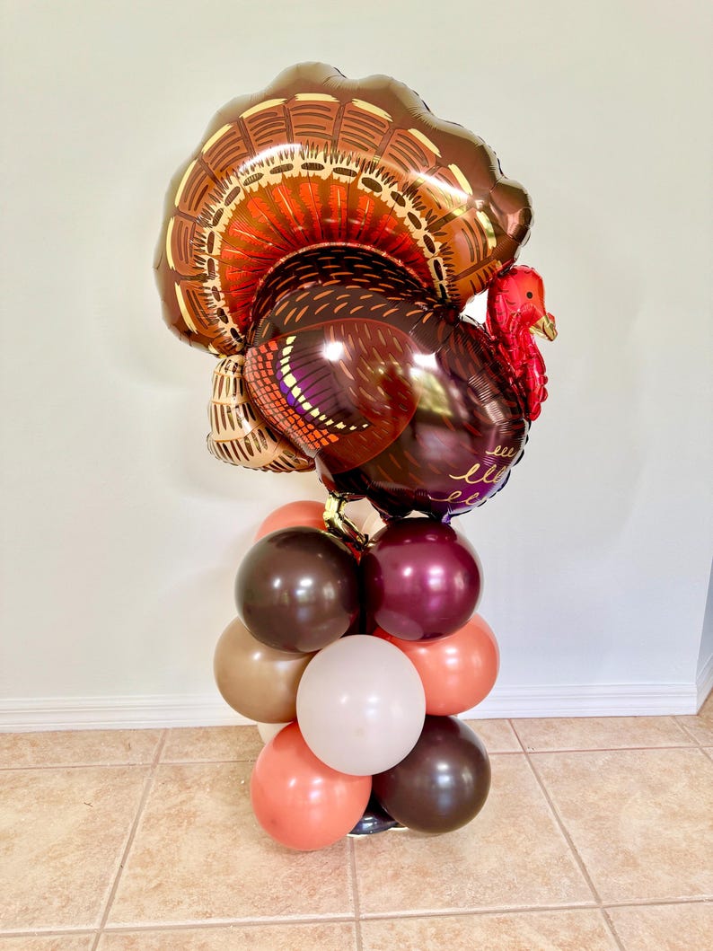May include: A large brown and red foil balloon turkey with a gold and brown balloon arch underneath. The balloon arch is made of brown, beige, and orange balloons.