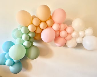 May include: A pastel rainbow balloon arch made of light blue, green, peach, pink, and white balloons. The balloons are arranged in a cascading arch shape.