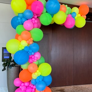 May include: A colourful balloon arch made of neon pink, green, blue, yellow, and orange balloons. The arch is arranged in a cascading pattern and is perfect for a party or event.