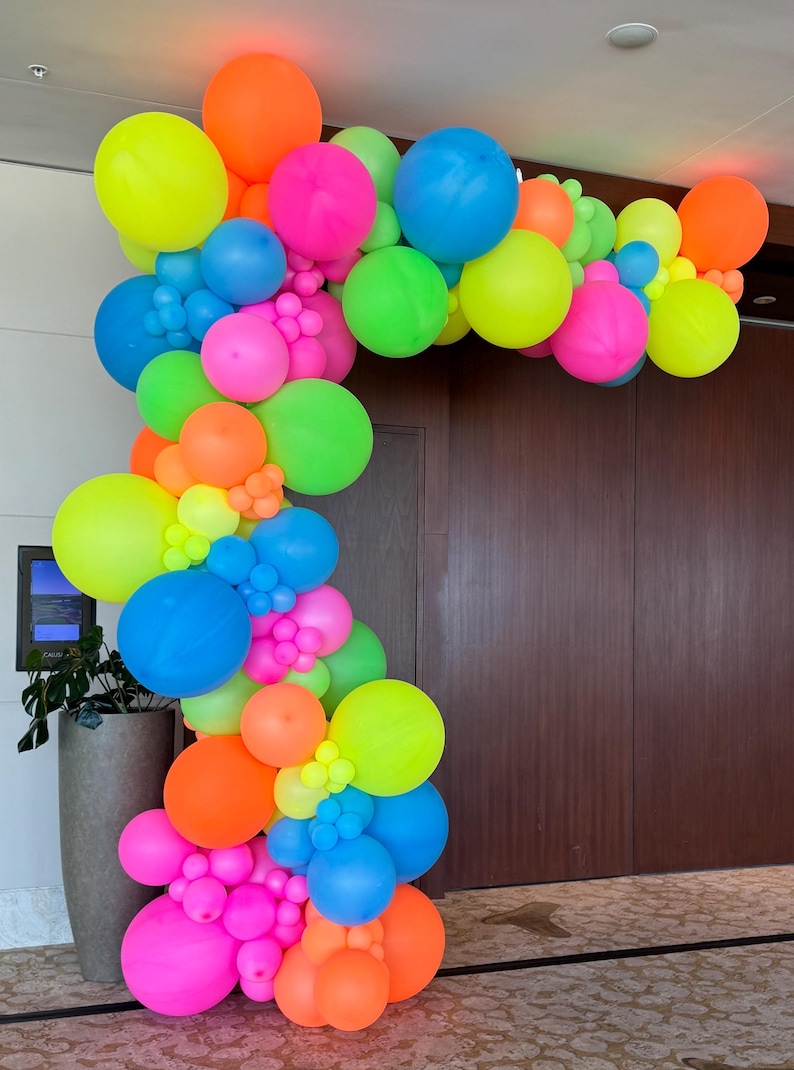 May include: A colourful balloon arch made of neon pink, green, blue, yellow, and orange balloons. The arch is arranged in a cascading pattern and is perfect for a party or event.