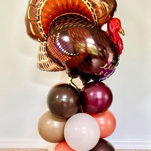 May include: A brown and gold turkey balloon with a red head and wattle, surrounded by a cluster of brown, burgundy, beige, and peach balloons.