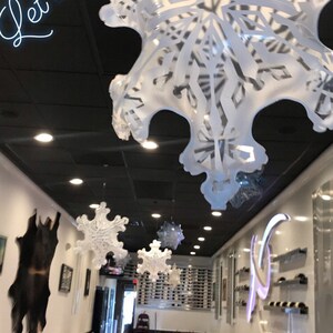 K&ouml;nnte beinhalten: Ein Restaurant-Interieur, das mit wei&szlig;en Schneeflocken-Papierdekorationen geschm&uuml;ckt ist, die von der Decke h&auml;ngen. Der Text "Let it snow" ist in wei&szlig;en Neonbuchstaben an der schwarzen Decke geschrieben. Das Restaurant hat wei&szlig;e Tischdecken und schwarze St&uuml;hle.