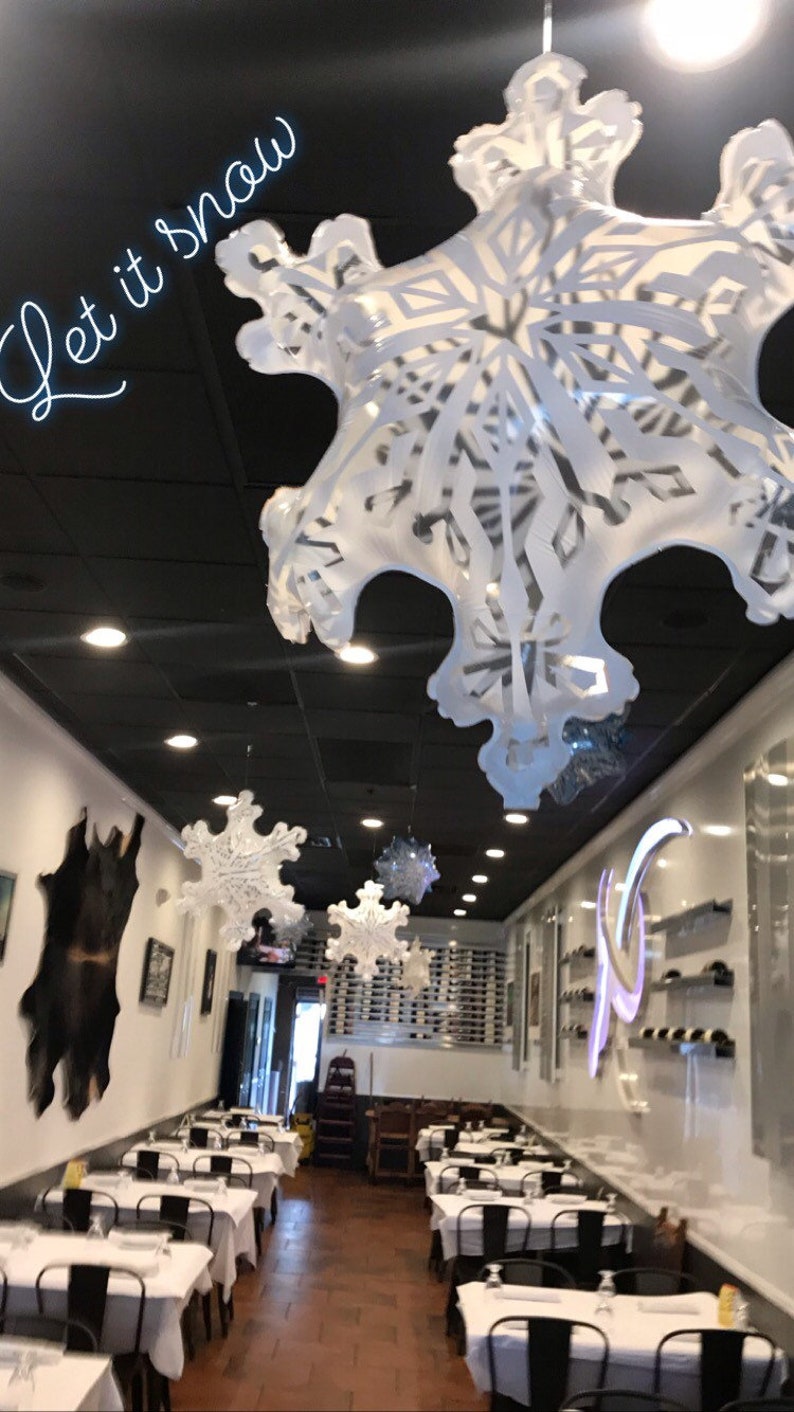 K&ouml;nnte beinhalten: Ein Restaurant-Interieur, das mit wei&szlig;en Schneeflocken-Papierdekorationen geschm&uuml;ckt ist, die von der Decke h&auml;ngen. Der Text "Let it snow" ist in wei&szlig;en Neonbuchstaben an der schwarzen Decke geschrieben. Das Restaurant hat wei&szlig;e Tischdecken und schwarze St&uuml;hle.