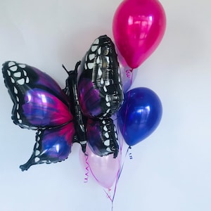 Pu&ograve; includere: Un gruppo di palloncini, tra cui un grande palloncino a forma di farfalla con ali viola e nere. Palloncini magenta, blu e rosa chiaro accompagnano la farfalla. I palloncini sono legati con un nastro viola, adatti per una festa.