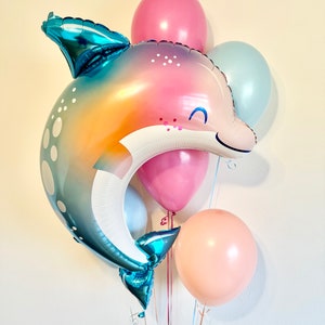 May include: A cluster of balloons featuring a large iridescent blue and pink dolphin balloon with a smiling face, a pink balloon, a light blue balloon, and a peach-colored balloon.