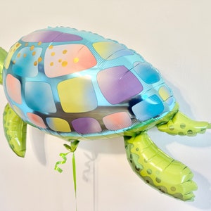 May include: A sea turtle-shaped balloon with a colorful design. The balloon's shell features blue, purple, yellow, and gold squares. The head and flippers are green. A green ribbon is attached to the balloon.