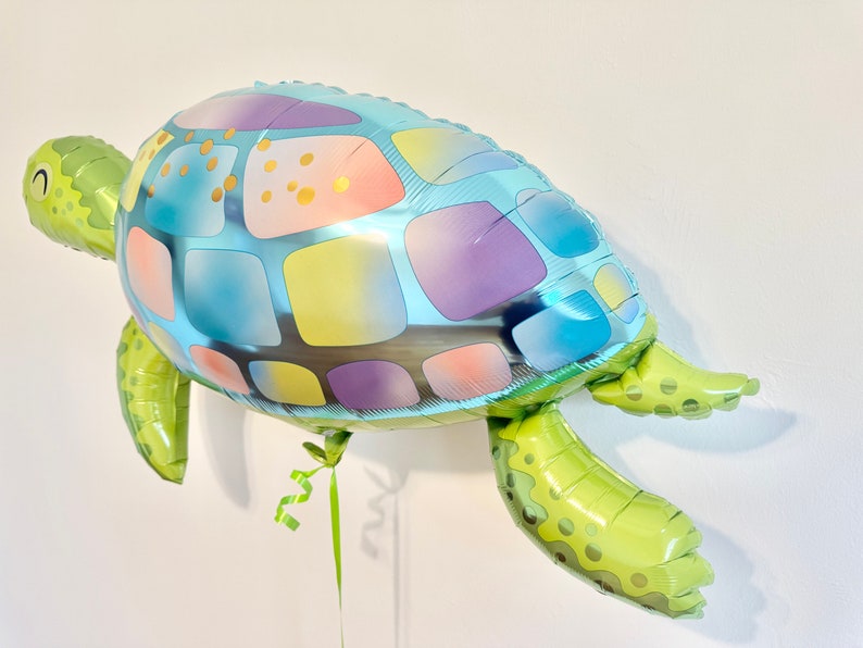 May include: A sea turtle-shaped balloon with a colorful design. The balloon's shell features blue, purple, yellow, and gold squares. The head and flippers are green. A green ribbon is attached to the balloon.