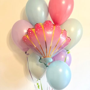 May include: A bouquet of pastel balloons with a large pink and gold seashell balloon in the center. The balloons are tied together with green and white ribbons.