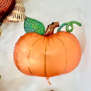 Puede incluir: Un globo grande de color naranja en forma de calabaza con una hoja verde con purpurina y un tallo marr&oacute;n. El globo est&aacute; atado a una cuerda.