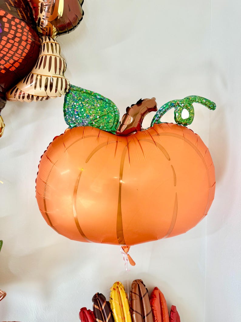 Puede incluir: Un globo grande de color naranja en forma de calabaza con una hoja verde con purpurina y un tallo marr&oacute;n. El globo est&aacute; atado a una cuerda.