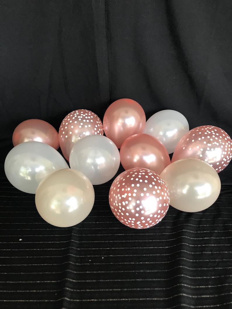 May include: A collection of party balloons in various colors, including white, rose gold, and a light peach. Some balloons have a polka dot pattern. The balloons are arranged on a black striped surface.