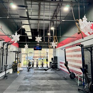 K&ouml;nnte beinhalten: Ein Indoor-Fitnessstudio mit schwarzen W&auml;nden, einer rot-wei&szlig; gestreiften amerikanischen Flagge und mehreren Trainingsger&auml;ten, darunter Ruderger&auml;te und eine Klimmzugstange. Au&szlig;erdem h&auml;ngen mehrere Schneeflocken-Dekorationen von der Decke.