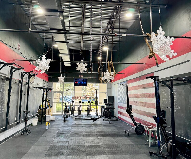 K&ouml;nnte beinhalten: Ein Indoor-Fitnessstudio mit schwarzen W&auml;nden, einer rot-wei&szlig; gestreiften amerikanischen Flagge und mehreren Trainingsger&auml;ten, darunter Ruderger&auml;te und eine Klimmzugstange. Au&szlig;erdem h&auml;ngen mehrere Schneeflocken-Dekorationen von der Decke.