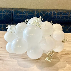 May include: A cluster of white balloons, some large and some small, arranged together. Delicate white flowers are interspersed among the balloons. The arrangement sits on a light-colored wooden surface.