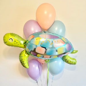 May include: A festive balloon bouquet featuring a large, smiling turtle-shaped foil balloon in shades of green, blue, and pastel colors. The turtle is surrounded by several round balloons in light pink, lavender, and blue hues, tied together with ribbons.