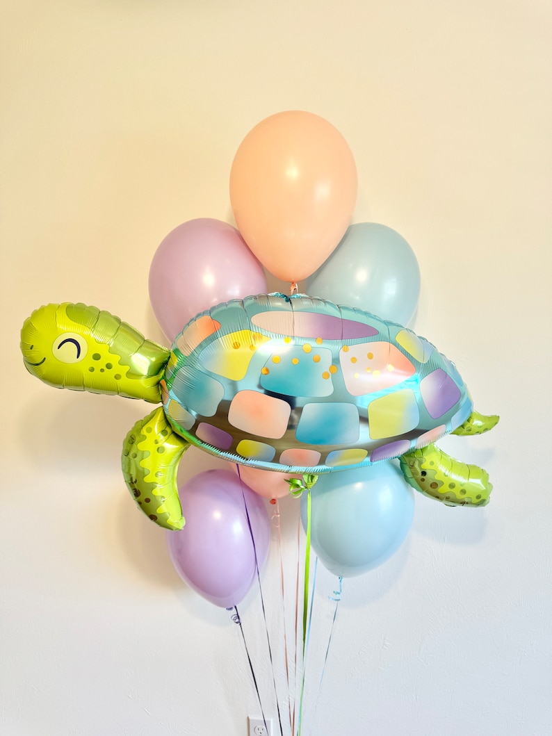 May include: A festive balloon bouquet featuring a large, smiling turtle-shaped foil balloon in shades of green, blue, and pastel colors. The turtle is surrounded by several round balloons in light pink, lavender, and blue hues, tied together with ribbons.
