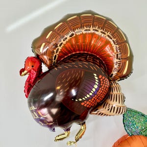 Puede incluir: Un globo de aluminio de color marr&oacute;n y dorado con forma de pavo con cabeza y barbilla rojas. El pavo tiene un dise&ntilde;o detallado de plumas en su cuerpo y cola.
