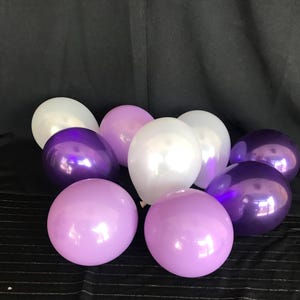 May include: A collection of nine balloons in shades of purple and white. The balloons are arranged on a black surface, with a dark background. The balloons have a shiny, reflective surface.