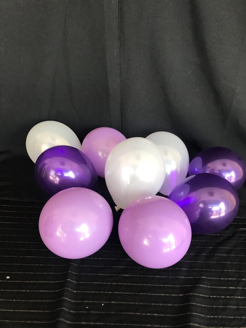 May include: A collection of nine balloons in shades of purple and white. The balloons are arranged on a black surface, with a dark background. The balloons have a shiny, reflective surface.