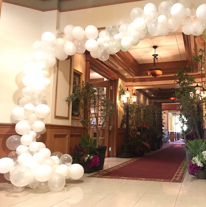 May include: A white balloon archway decorates an entrance to a room with a red carpet and potted plants.