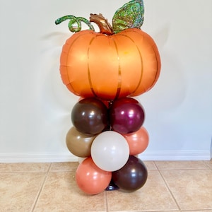 May include: A balloon centerpiece featuring a large orange pumpkin balloon with a green stem and leaves. The pumpkin balloon is on top of a stack of smaller balloons in shades of brown, burgundy, and cream.