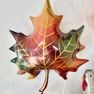 Puede incluir: Un gran globo met&aacute;lico de color dorado, rojo y verde en forma de hoja. El globo tiene un acabado brillante y tiene la forma de una hoja de arce.
