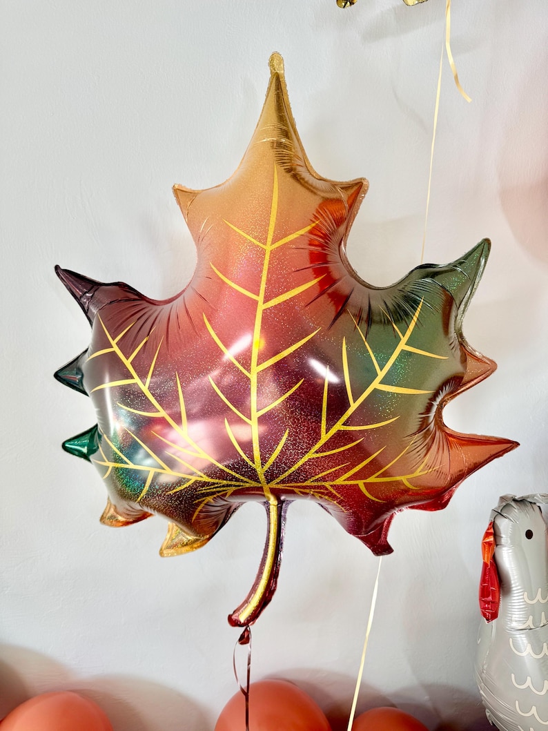 Puede incluir: Un gran globo met&aacute;lico de color dorado, rojo y verde en forma de hoja. El globo tiene un acabado brillante y tiene la forma de una hoja de arce.