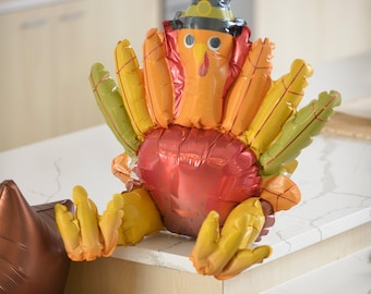 May include: A red, orange, and yellow foil balloon turkey with a brown pilgrim hat. The turkey is sitting on a white countertop.
