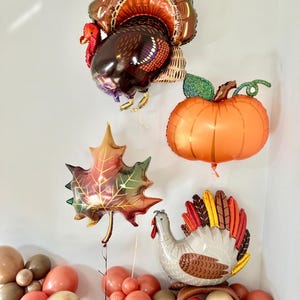 Puede incluir: Un arreglo de globos con tem&aacute;tica de Acci&oacute;n de Gracias con un globo de pavo marr&oacute;n y naranja, un globo de calabaza naranja grande, un globo de hoja roja y dorada y un globo de pavo gris y marr&oacute;n. Los globos est&aacute;n rodeados de una variedad de globos m&aacute;s peque&ntilde;os en tonos marrones, dorados y rojos.