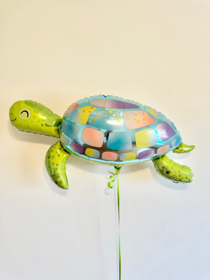 May include: A large, foil turtle-shaped balloon. The turtle has a green head, flippers, and tail, with a smiling face. The shell is a patchwork of blue, pink, yellow, and purple. A green ribbon hangs from the bottom.