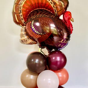 May include: A large brown and red turkey balloon with a gold and brown tail, standing on a stack of brown, beige, and red balloons. The turkey balloon has the word "bless" written on it.