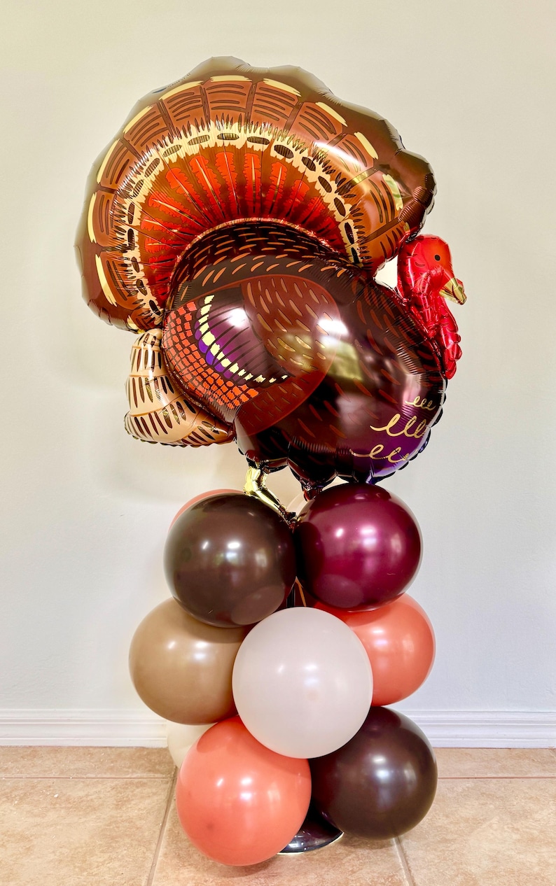 May include: A large brown and red turkey balloon with a gold and brown tail, standing on a stack of brown, beige, and red balloons. The turkey balloon has the word "bless" written on it.