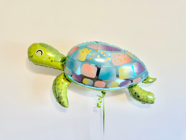 May include: A turtle-shaped foil balloon in shades of green, blue, and pastel colors. The balloon has a smiling face and is designed to resemble a sea turtle. It is suspended by a green ribbon.
