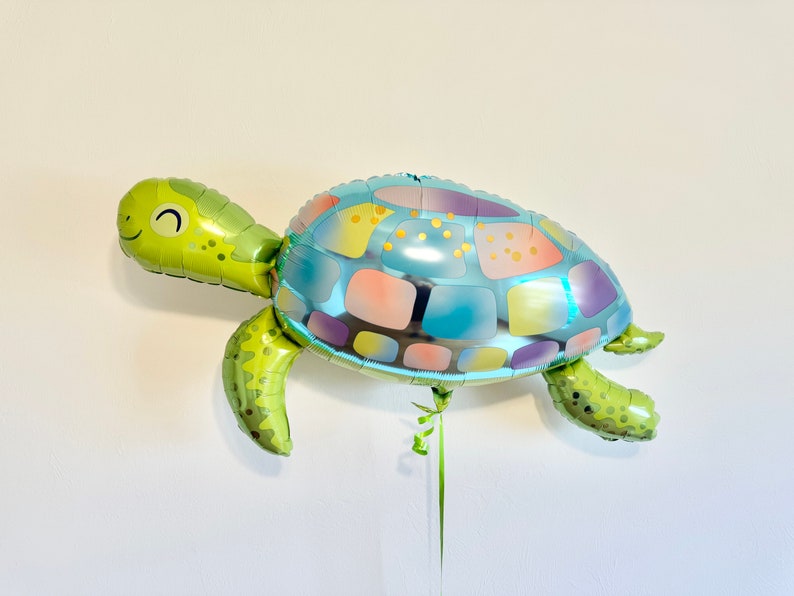 May include: A turtle-shaped foil balloon. The balloon features a green head and limbs, and a shell with a blue, purple, and yellow pattern. The balloon has a smiling face and is suitable for a birthday party or other celebration.