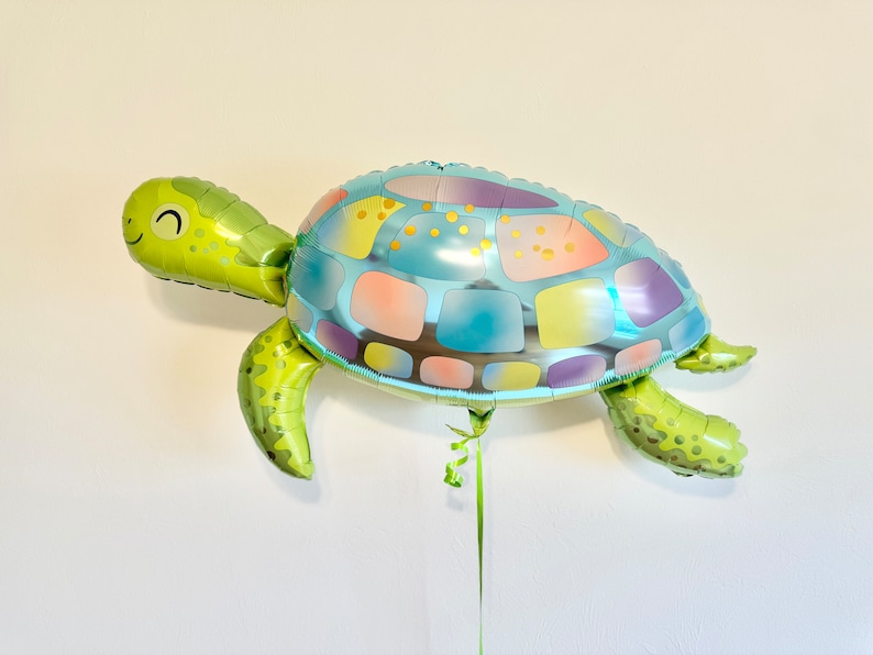 May include: A green and blue turtle-shaped balloon. The balloon has a smiling face and a shell with pastel-colored panels. The balloon is suspended by a green ribbon, perfect for a birthday party or ocean-themed event.