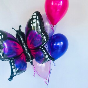 Pu&ograve; includere: Un gruppo di palloncini, tra cui un grande palloncino a forma di farfalla con ali nere e viola. Ulteriori palloncini sono in tonalit&agrave; di rosa, viola e blu. Nastri e serpentini sono attaccati, creando una composizione festosa.