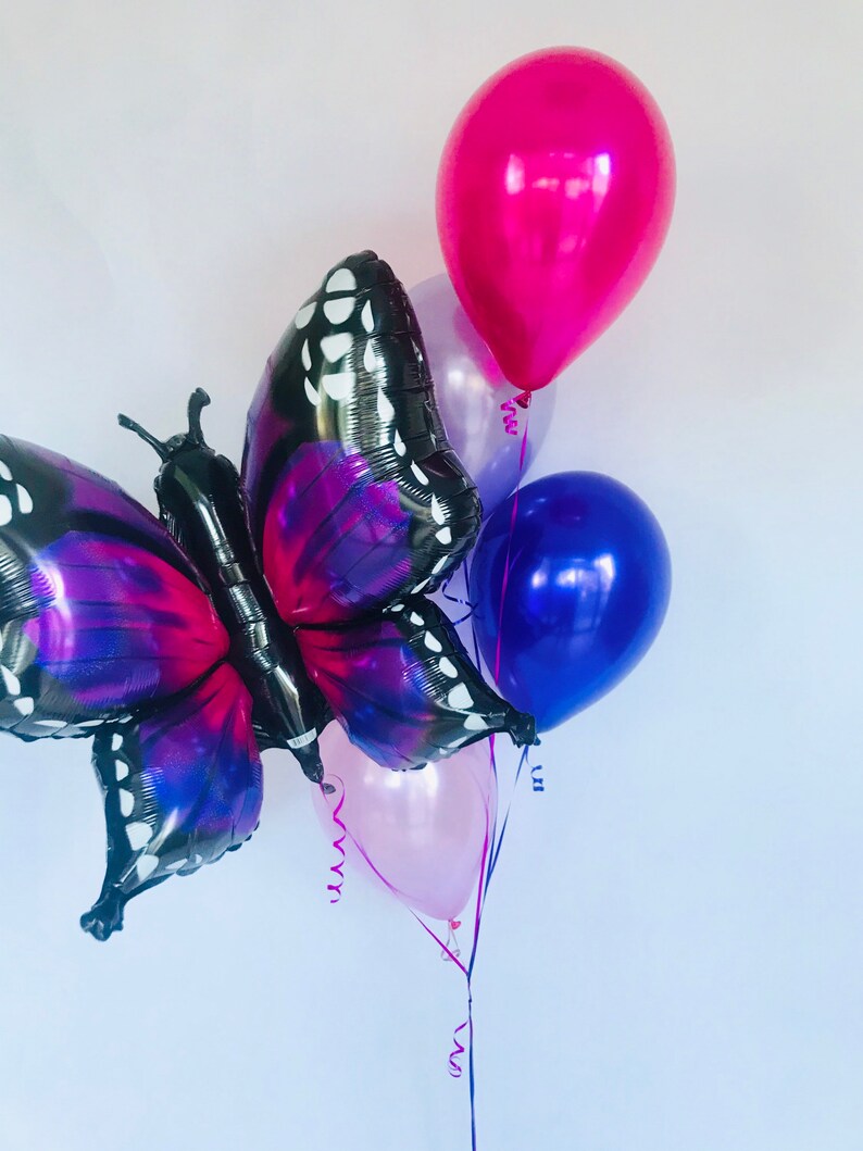 Pu&ograve; includere: Un gruppo di palloncini, tra cui un grande palloncino a forma di farfalla con ali nere e viola. Ulteriori palloncini sono in tonalit&agrave; di rosa, viola e blu. Nastri e serpentini sono attaccati, creando una composizione festosa.