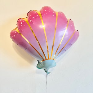 May include: A seashell-shaped balloon in shades of pink and purple, with gold vertical stripes and white polka dots. The balloon has a light blue base and a silver string, perfect for a mermaid or ocean-themed party.