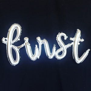 May include: Silver foil balloons spelling out the word "first" against a black background.